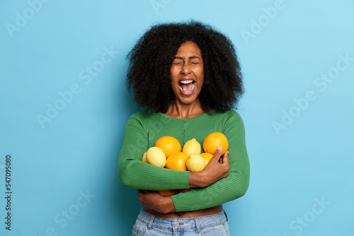 Indoor shot of emotional curly girl holding fresh oranges and lemons fruits  keeps mouth widely opened  dressed in casual cloth