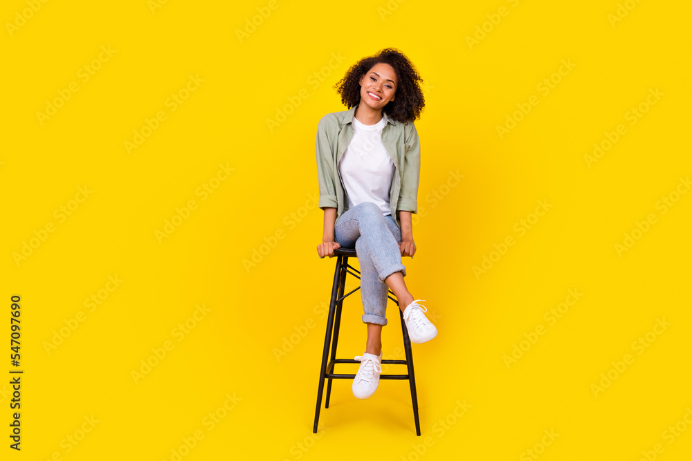 Full size photo of cheery young woman curly hair sit chair have rest chatting wear trendy khaki outfit isolated on yellow color background Full size photo of cheery young woman curly hair sit chair have rest chatting wear trendy khaki outfit isolated on yellow color background