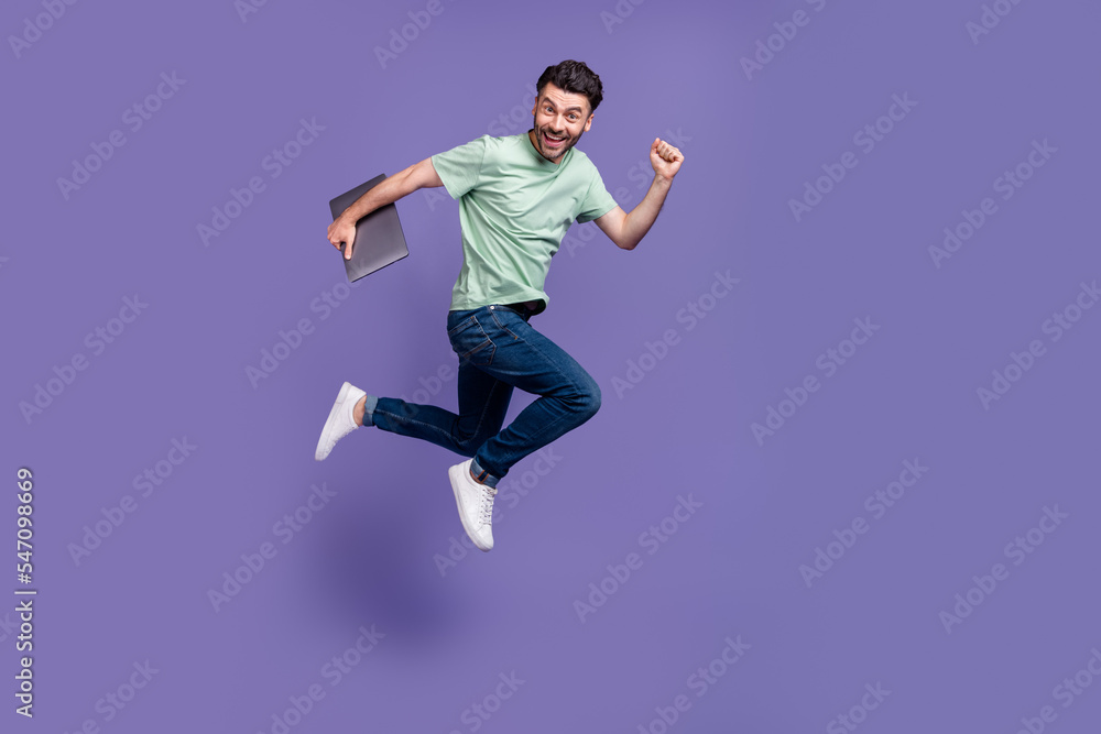 Full size photo of handsome young man running fast jump hold laptop dressed stylish gray clothes isolated on purple color background Full size photo of handsome young man running fast jump hold laptop dressed stylish gray clothes isolated on purple color background