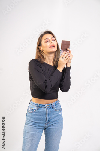A beautiful brown-haired girl holds a chocolate bar on a white background. Young brunette woman wearing a black blouse and blue jeans