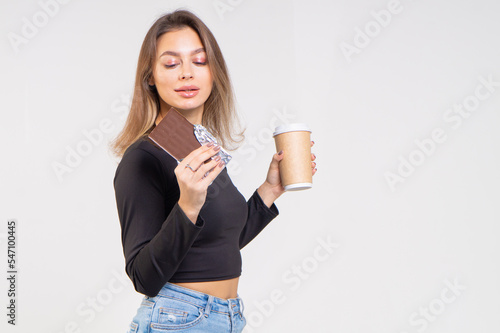 A beautiful brown-haired girl holds a paper cup with coffee and chocolate bar on a white background. Young brunette woman wearing a black blouse and blue jeans