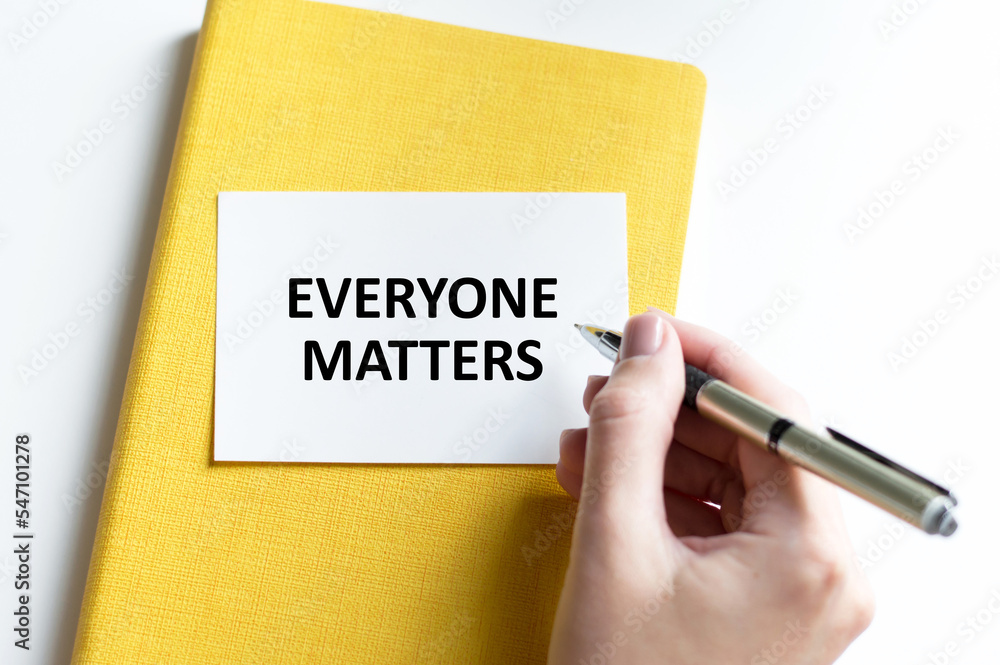 EVERYONE MATTERS text on a card on a yellow notebook on a light background next to a person's hand with a pen EVERYONE MATTERS text on a card on a yellow notebook on a light background next to a person's hand with a pen