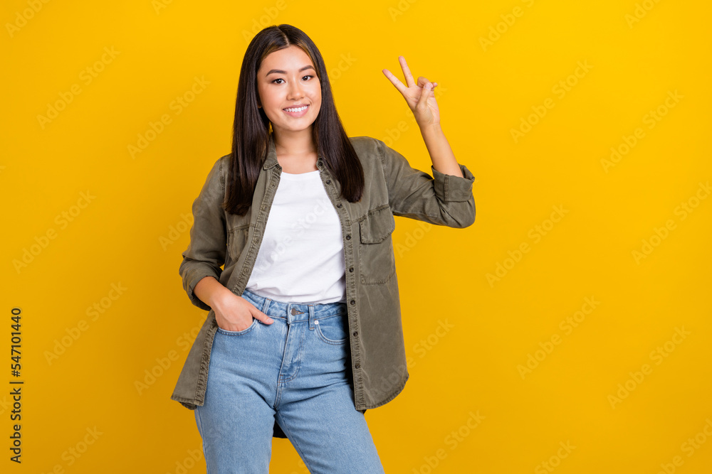 Portrait of pleasant gorgeous girl long hairdo wear khaki shirt hold arm in pocket showing v-sign isolated on yellow color background Portrait of pleasant gorgeous girl long hairdo wear khaki shirt hold arm in pocket showing v-sign isolated on yellow color background