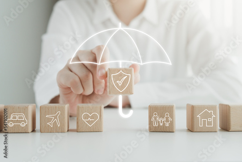 Insurance concept. Protection against a possible eventuality. House, Car, Family, travel and Health on wooden block and Hand holding security icon cover by umbrella icon for life assurance concept.