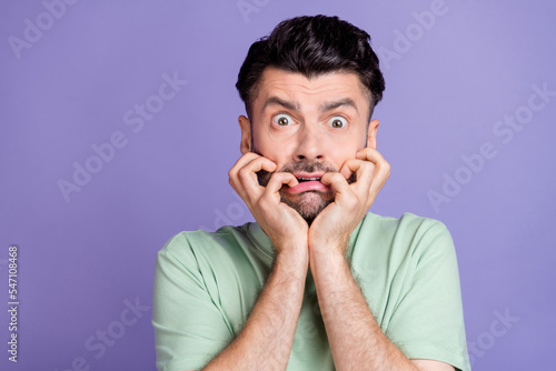 Photo of afraid fearful guy with brunet hairstyle stubble wear t-shirt hands on cheeks scared staring isolated on purple color background