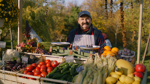 Konstfotografi Elderly farmer stands at the stall with fresh colorful fruits and vegetables, looks at camera