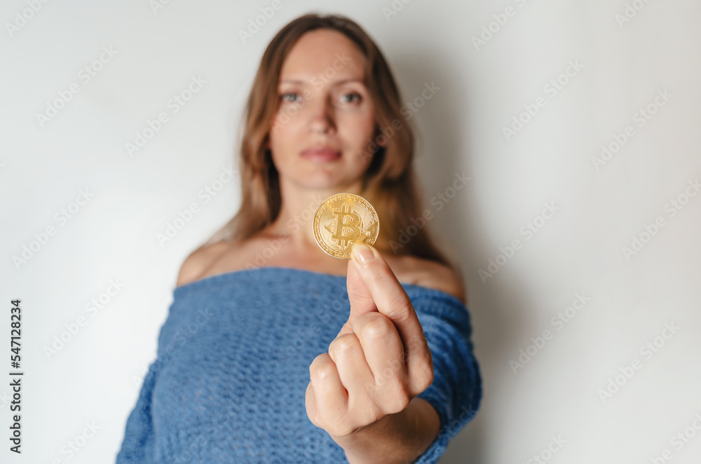 Joyful woman standing holding bitcoin paying attention to new digital cryptocurrency wearing blue sweater. Indoor studio shot isolated on white background.