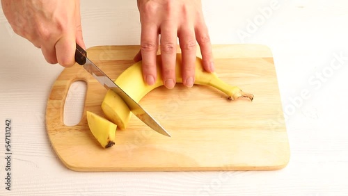 Male hand cuts fresh banana with a knife on wooden kitchen board. Side view, close up.