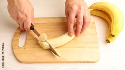 Male hand cuts fresh peeled banana with a knife on wooden kitchen board. Side view, close up.