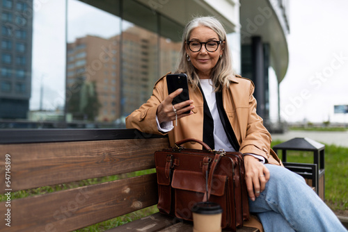 an older woman with a mobile phone sits on a bench outside