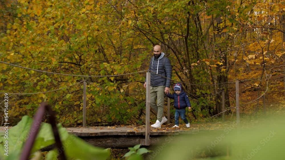 A man and his son, holding hands, walk across a bridge in an autumn forest or park. A man takes his little son in his arms and circles him on a suspension bridge. A man and his son, holding hands, walk across a bridge in an autumn forest or park. A man takes his little son in his arms and circles him on a suspension bridge.