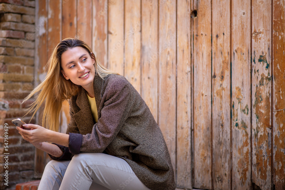 Smiling woman holding mobile phone and looking away while sitting outdoors against old wooden door Smiling woman holding mobile phone and looking away while sitting outdoors against old wooden door