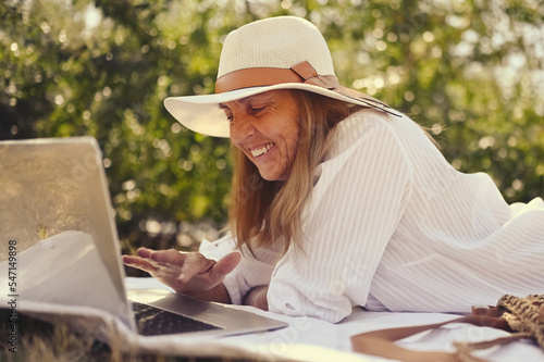 Senior happy woman working or having video call with laptop in summer outdoors. Old elegant lady in straw hat and bag sitting on grass during picnic at countryside. Active retired people concept