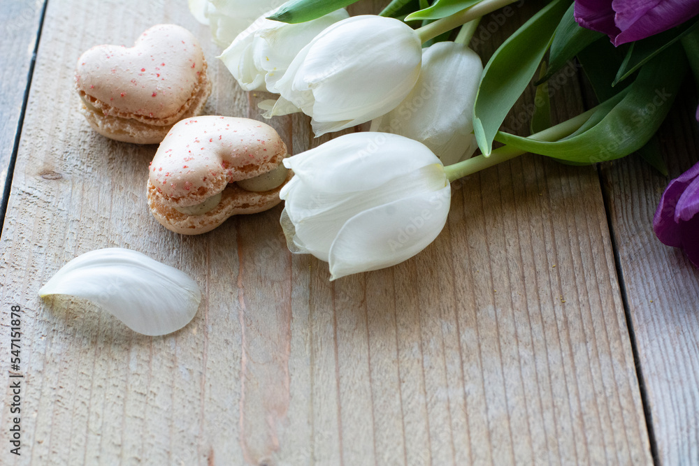 Two tasty creamy macarons are lying on the wooden table with white tulips. Two macarons in form of the heart as 8. Dessert is on the light wooden table