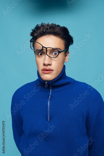 a handsome, attractive man stands on a light blue background in a blue zip-up sweater with black-rimmed glasses turned upside down on his face and looks in surprise at the camera with his mouth open