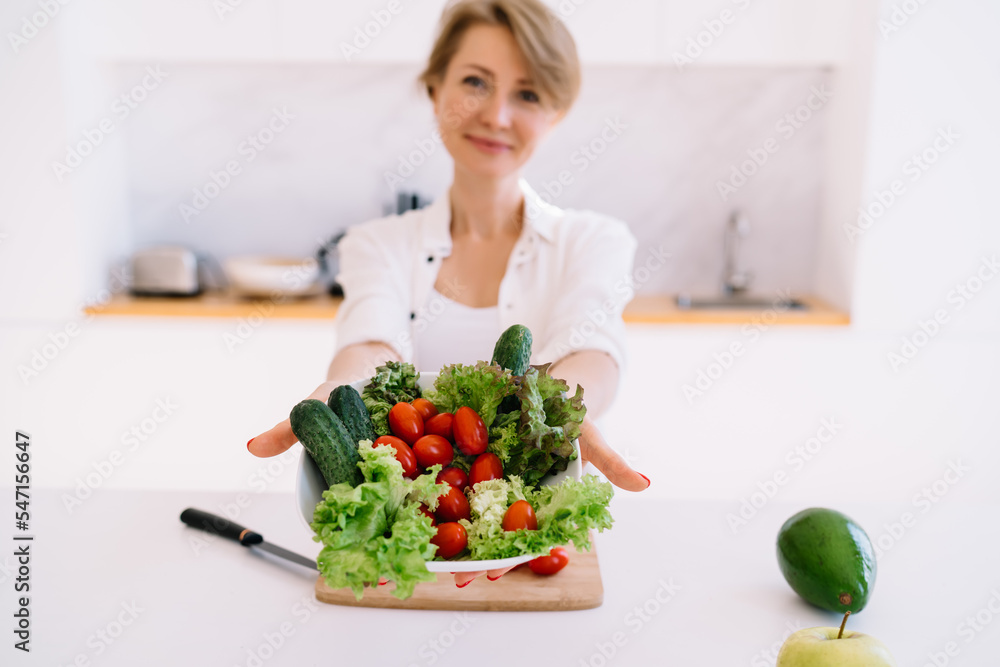 Smiling adult woman showing fresh vegetables in bowl on table at home Smiling adult woman showing fresh vegetables in bowl on table at home