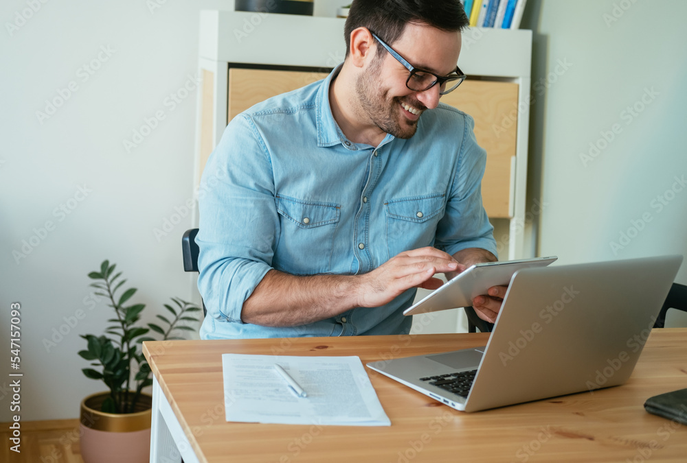 Smiling businessman typing report on his tablet while working at home office desk with a laptop computer, paper and pen Smiling businessman typing report on his tablet while working at home office desk with a laptop computer, paper and pen