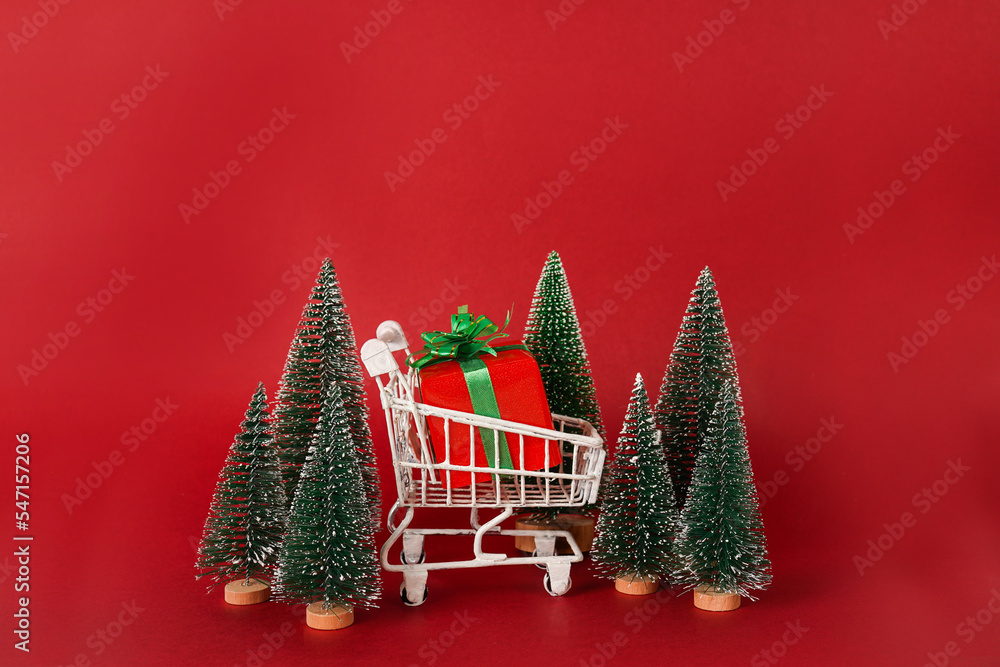 Miniature shopping cart with Christmas trees and gift box on red background with copy space. Christmas shopping concept Miniature shopping cart with Christmas trees and gift box on red background with copy space. Christmas shopping concept