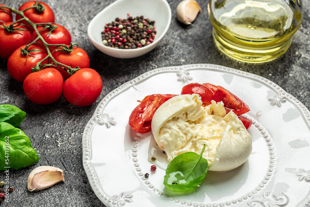 Salad with creamy Italian Burrata Cheese Served with Olive Oil, Basil Leaves and cherry tomatoes on white plate on a dark background. Restaurant menu, dieting, cookbook recipe top view