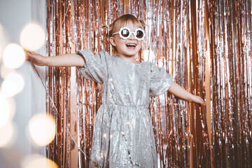 Happy little stylish girl in shiny dress having fun. Festive background with foil curtain decorations for kids birthday or fancy dress party, disco music or New Year.