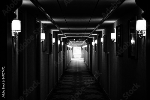 A hotel corridor in the dark with a light at the end of the hallway. Spooky black and white image.