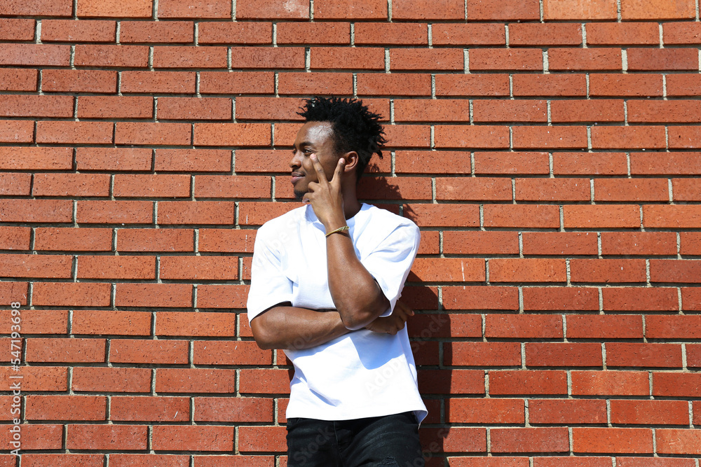 Young black man with dread fade haircut standing on a street and showing a peace gesture. Portrait of a guy in casual outfit posing over the red brick wall background with a copy space for text. Young black man with dread fade haircut standing on a street and showing a peace gesture. Portrait of a guy in casual outfit posing over the red brick wall background with a copy space for text.