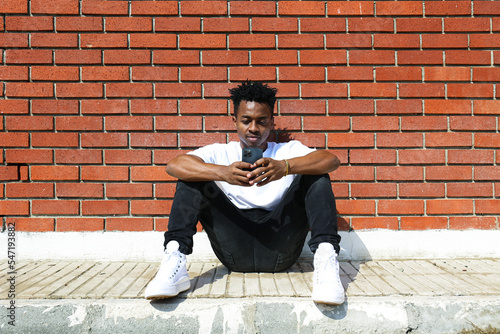 Young black man on the street sitting on the curb with his phone and texting. Portrait of a guy wearing a casual outfit leaning on the red brick wall with copy space for text. Background. © Evrymmnt