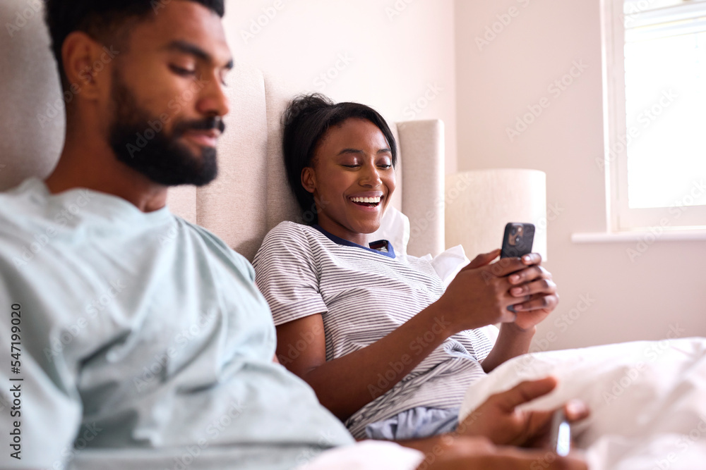 Young Couple At Home Both Looking At Mobile Phones In Bed Together Young Couple At Home Both Looking At Mobile Phones In Bed Together
