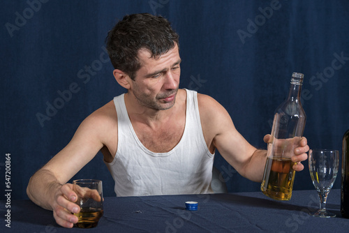 Lonely drunk man in white home shirt sits at table and looks at bottle of alcohol that lies in his hand. In other hand he holds glass of vodka. Alcoholism as disease. photo