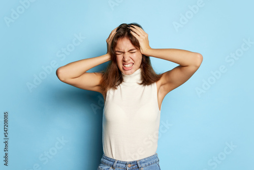 Portrait of young beautiful woman posing with hands on head isolated over blue studio background. Irritation