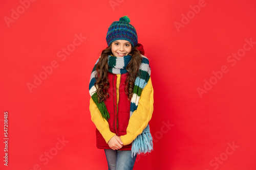 cheerful kid in knitted winter hat and scarf on red background, cold winter