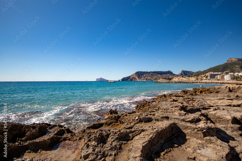 Panorámica de la zona rocosa de Calpe con las olas  del mar Mediterráneo rompiendo en sus piedras bajo un precioso cielo azul soleado en un día de otoño.