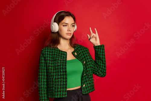 Portrait of young beautiful woman posing, listening to rock music in headphones isolated over red studio background