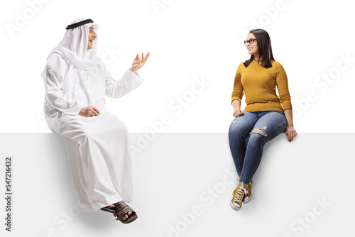 Arab man in an ethnic robe sitting on a blank panel and talking to a casual young woman