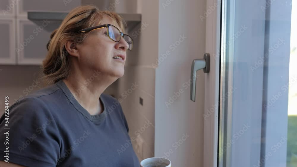 Mature Woman Holding a Cup of Tea or Coffee While Standing in Front of Windows During the Day Mature Woman Holding a Cup of Tea or Coffee While Standing in Front of Windows During the Day