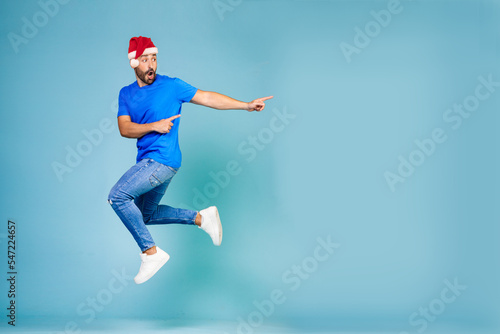 Full length portrait of a cheerful young man jumping in a blue t-shirt and a New Year's hat, pointing his finger to the side on a blue background