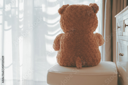 Brown teddy bear sitting alone on white cotton chair with sunlight from the door or window in front of house, Cinematic tone style vintage, Lonely and waiting for lover or family concept. photo
