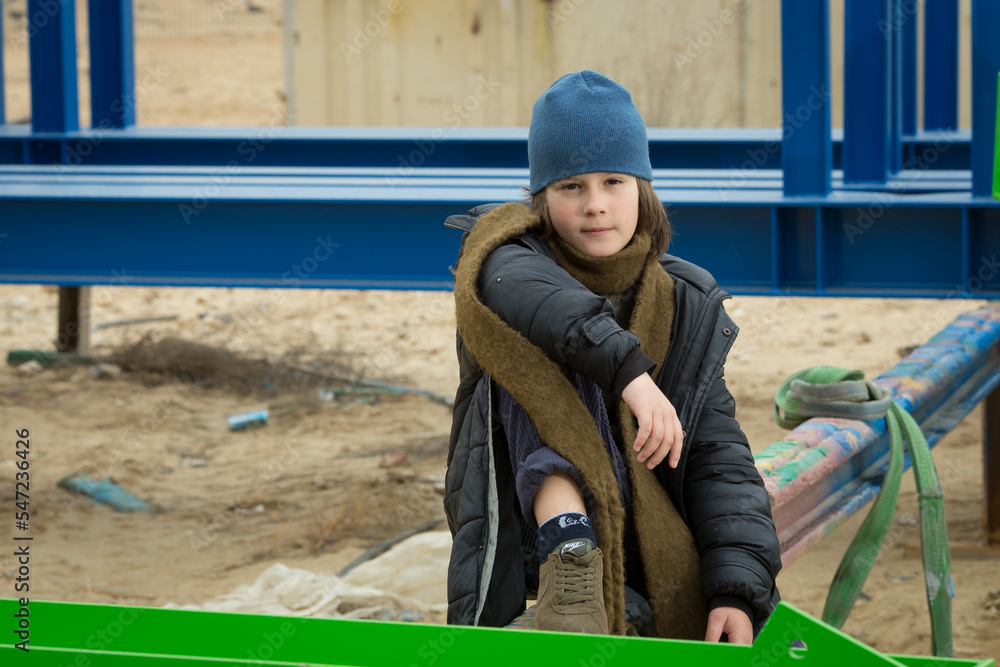 cool stylish caucasian boy in a coat and hat with a scarf posing coolly plays and walks through pipes in an industrial area among green yellow blue metal structures against a cloudy sky on a rainy day cool stylish caucasian boy in a coat and hat with a scarf posing coolly plays and walks through pipes in an industrial area among green yellow blue metal structures against a cloudy sky on a rainy day