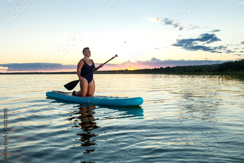 A woman with a short Mohawk haircut in a swimsuit on a SUP board with a paddle floats on the water.