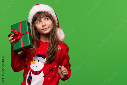 An emotional girl in a knitted sweater and a Santa hat holds a gift in her hands and looks up dreamily. Guess the surprise. Merry Christmas and Happy New Year 2023