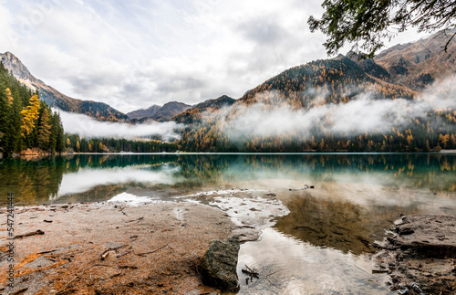 Panoramic view of Lake Anterselva in Trentino Alto Adige, Italy. Scenic alpine landscape with autumn pine forests, misty clouds over the turquoise water and reflection of the Dolomite mountains.