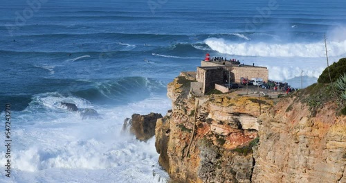 People watching the big giant waves crashing near the Fort of Nazare Lighthouse in Nazare, Portugal. Biggest waves in the world. Touristic destination for surfing.