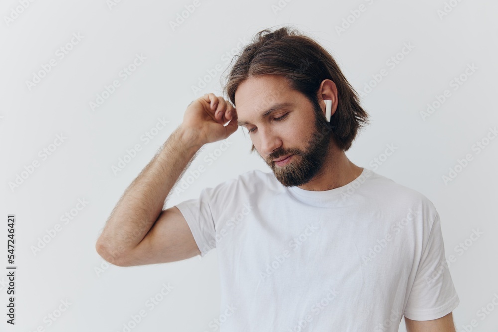 A man with a beard and long hair in a white T-shirt and blue jeans stands against a white wall, leaning against it and listening to music with wireless white headphones, staring thoughtfully A man with a beard and long hair in a white T-shirt and blue jeans stands against a white wall, leaning against it and listening to music with wireless white headphones, staring thoughtfully