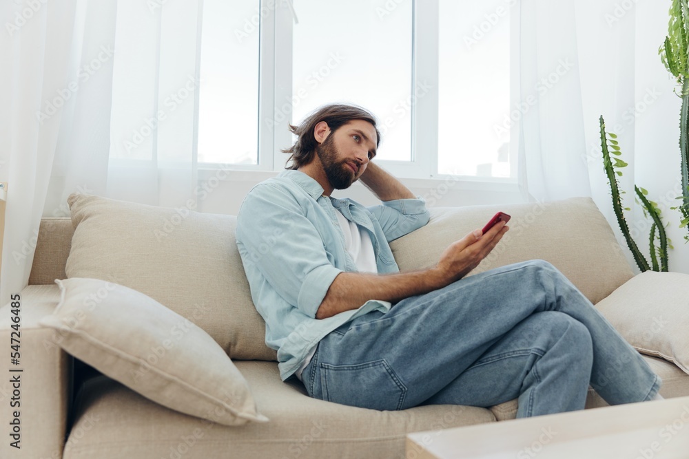A man with a beard sits on the couch during the day and smiles at home and looks at his phone relaxing on his day off, life online on his phone texting with friends A man with a beard sits on the couch during the day and smiles at home and looks at his phone relaxing on his day off, life online on his phone texting with friends