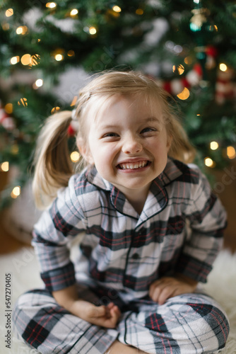 Little blonde girl with pigtails wearing pajamas and sitting in front of a Christmas tree smiling