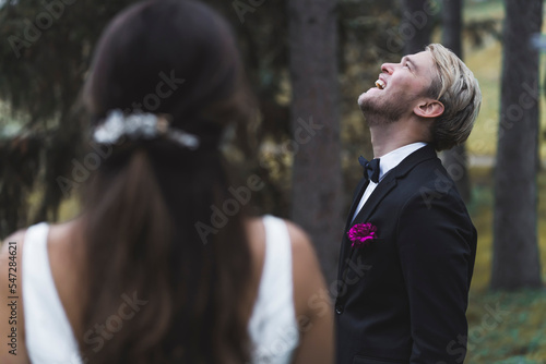 Focus on blond bearded laughing Scandinavian groom in elegant black tuxedo with butterfly tie and magenta-colored flower with his head upwards. Blurred back view of unrecognizable dark-hair bride in