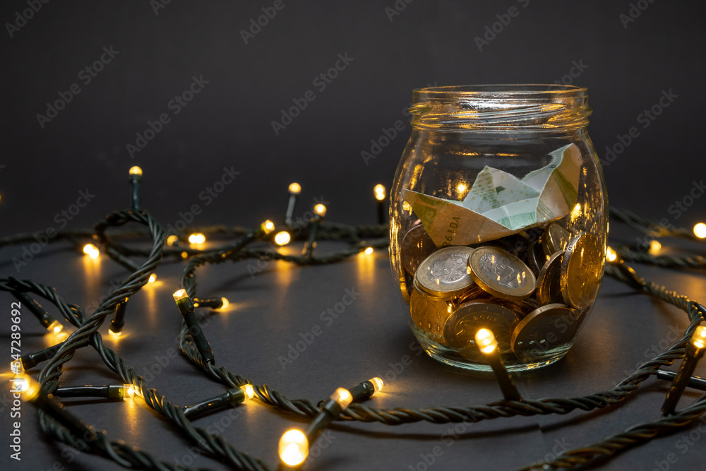 Glass jar with euro coins and paper boat on a dark background with small led lights. Euro pocket savings coin concept.