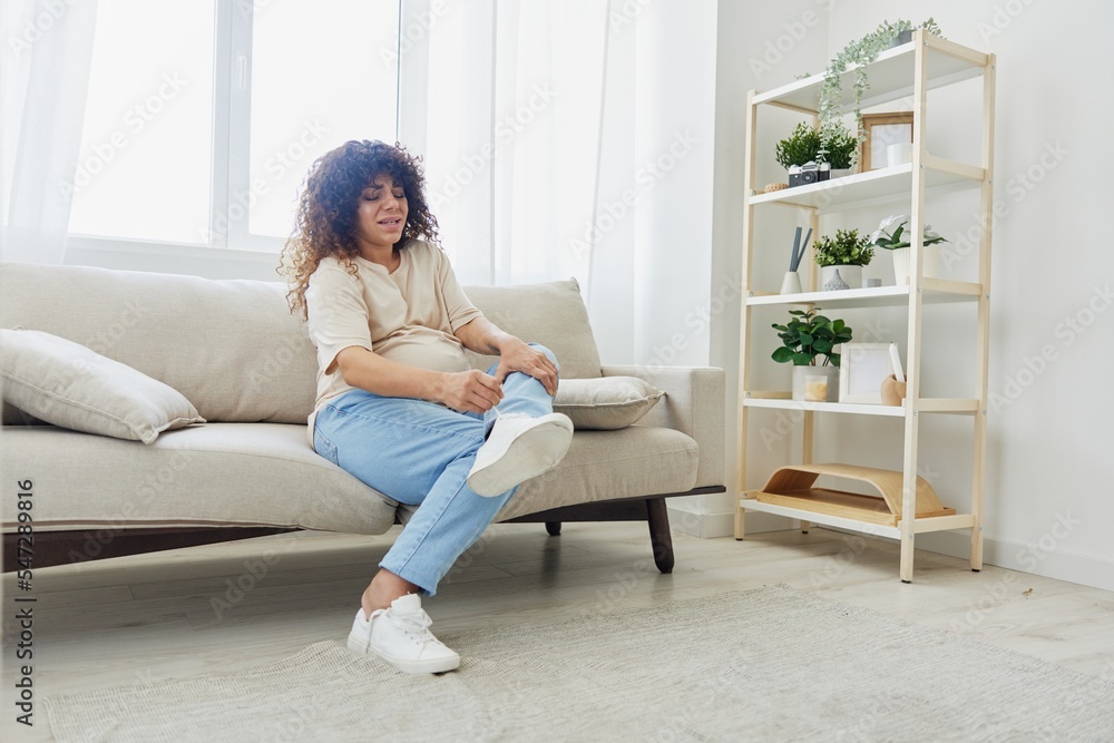 Pregnant woman sitting on sofa at home and putting on pregnancy-friendly shoes with problem due to belly, headache Pregnant woman sitting on sofa at home and putting on pregnancy-friendly shoes with problem due to belly, headache