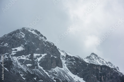 Awesome landscape with high snowy mountain peaked top with sharp rocks in cloudy sky. Dramatic view to snow mountain pointed peak in rainy weather. Atmospheric scenery with white snow on black rocks.