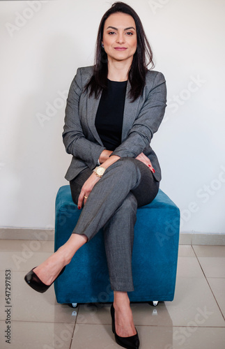 A mulher empreendedora em uma sessão de fotos corporativa em pose sentada no banco e sorridente vestida elegantemente com paletó e com mãos delicadas